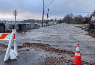 Photo of a flooded roadway with two orange cones and a barrier in the foreground and the bay in the background
